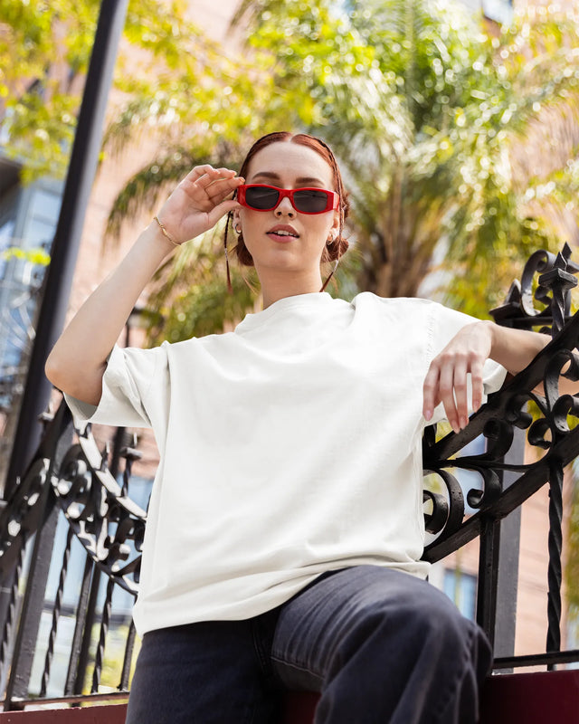 Woman wearing red sunglasses and a white t-shirt sitting outdoors with greenery in the background