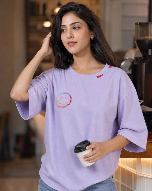Woman wearing a light lavender t-shirt with a colorful logo, holding a coffee cup in a casual setting.