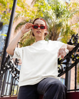 Woman wearing red sunglasses and a white t-shirt sitting outdoors with greenery in the background