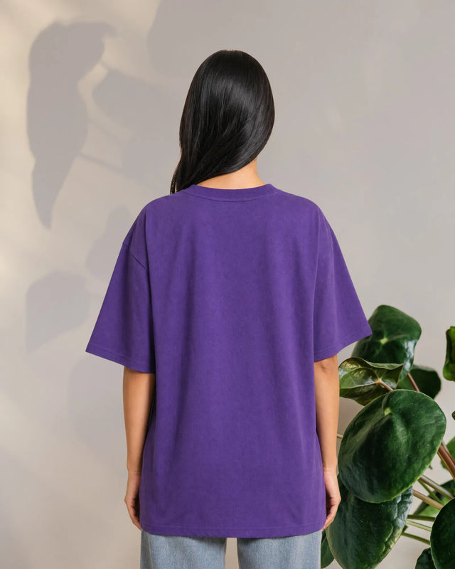 Person wearing a purple t-shirt standing against a neutral background with a plant to the side.