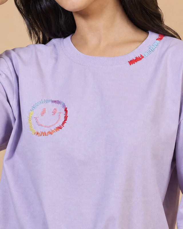 Person wearing a light lavender t-shirt with a colorful smiley face design on a beige background