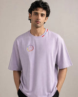 Man wearing a light lavender t-shirt with a colorful logo on a plain background